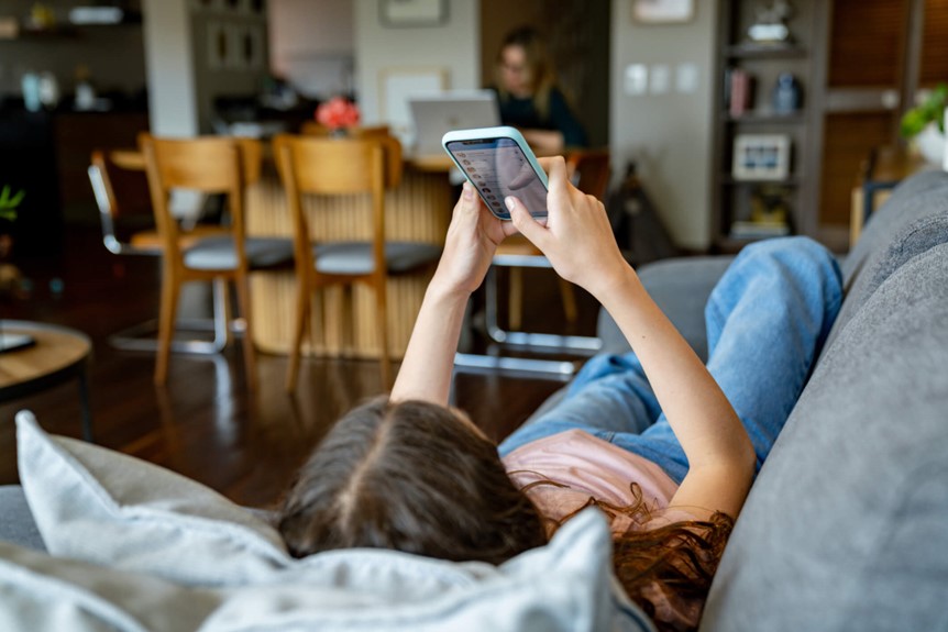 Person lying on a gray couch in a living room, holding a smartphone above while looking at the screen