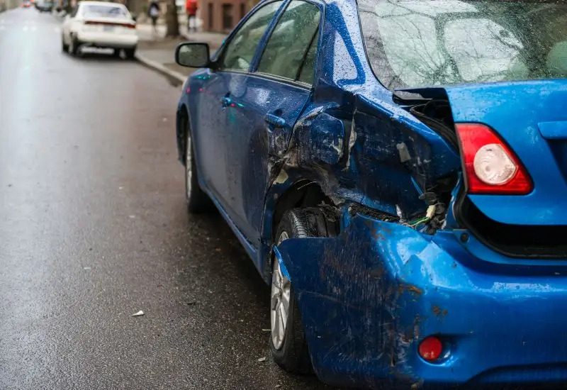 Blue car with significant rear-side damage from a hit-and-run accident on wet road