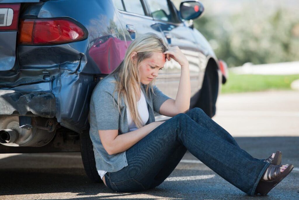 Person sitting on the ground next to a car with rear-end damage after an accident