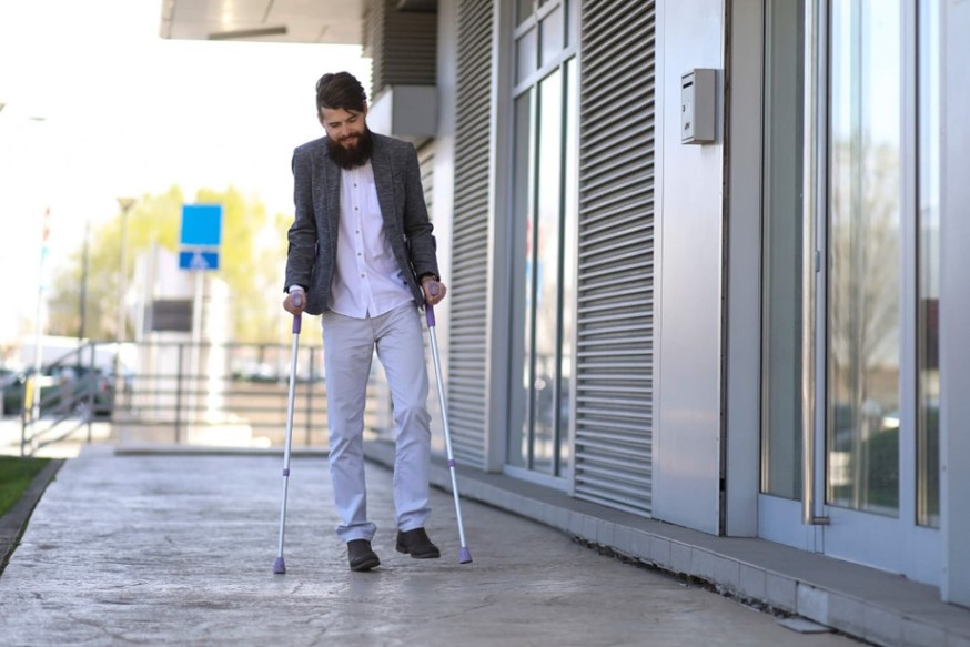 A person walking on an outdoor sidewalk using forearm crutches beside a Houston, TX, building